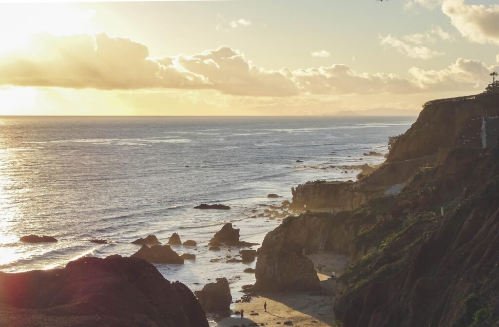 Sunlit coastline with rocky cliffs on the right, a small sandy beach, and a calm sea at sunset, with a few people strolling.